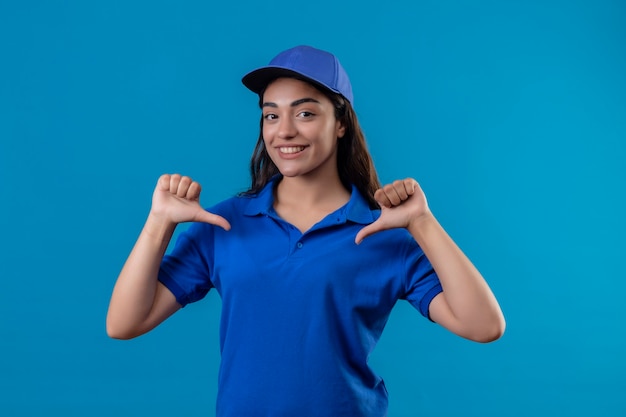 Jovem entregadora de uniforme azul e boné parecendo confiante apontando para si mesma com o polegar satisfeita e orgulhosa de pé sobre um fundo azul