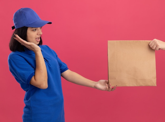 Jovem entregadora de uniforme azul e boné se sentindo animada ao receber um pacote de papel em pé sobre a parede rosa