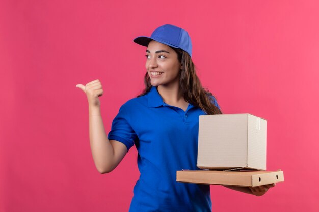 Jovem entregadora de uniforme azul e boné segurando caixas de papelão apontando com o dedo para o lado sorrindo alegremente feliz e positiva olhando de lado em pé sobre um fundo rosa