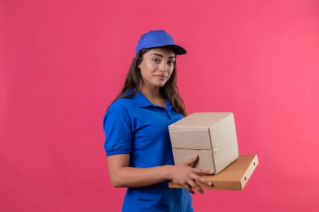 Jovem entregadora de uniforme azul e boné segurando caixas de papelão sorrindo, confiante, positiva e feliz em pé sobre um fundo rosa