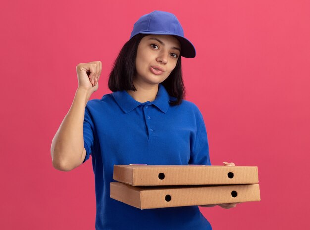 Jovem entregadora de uniforme azul e boné segurando caixas de pizza com os punhos cerrados, parecendo confiante em pé sobre a parede rosa