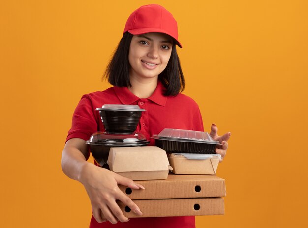 Jovem entregadora de uniforme azul e boné segurando caixas de pizza e pacote de comida sorrindo feliz e positiva em pé sobre a parede laranja