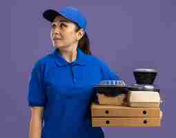 Foto grátis jovem entregadora de uniforme azul e boné segurando caixas de pizza e pacotes de comida olhando para o lado com um sorriso no rosto em pé sobre a parede roxa