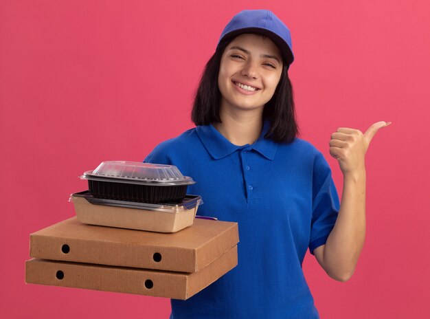 Jovem entregadora de uniforme azul e boné segurando caixas de pizza e um pacote de comida, sorrindo, apontando com o polegar para o lado em pé sobre a parede rosa