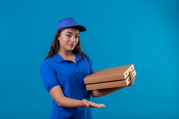 Jovem entregadora de uniforme azul e boné segurando caixas de pizza, olhando para a câmera, sorrindo confiante, feliz e positiva em pé sobre o fundo azul