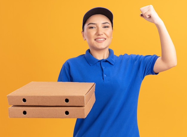 Jovem entregadora de uniforme azul e boné segurando caixas de pizza, olhando para a frente, sorrindo confiante e erguendo o punho como uma vencedora em pé sobre uma parede laranja