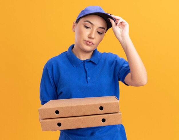 Jovem entregadora de uniforme azul e boné segurando caixas de pizza, olhando para elas se confundindo tocando seu boné em pé sobre a parede laranja