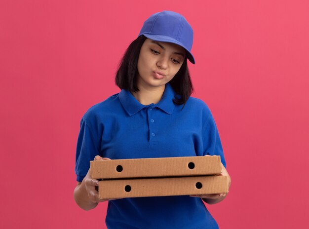 Jovem entregadora de uniforme azul e boné segurando caixas de pizza parecendo descontente com a expressão triste em pé sobre a parede rosa