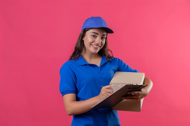 Jovem entregadora de uniforme azul e boné segurando o pacote da caixa e a prancheta escrevendo algo olhando para a câmera sorrindo alegremente feliz e positiva em pé sobre o fundo rosa