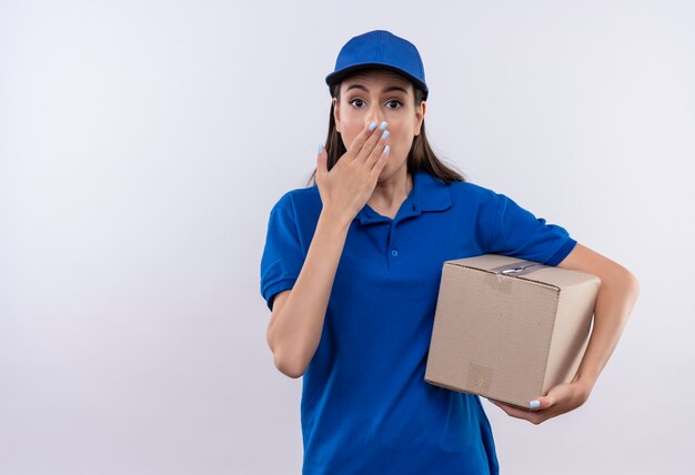 Jovem entregadora de uniforme azul e boné segurando um pacote de caixa chocada cobrindo a boca com a mão