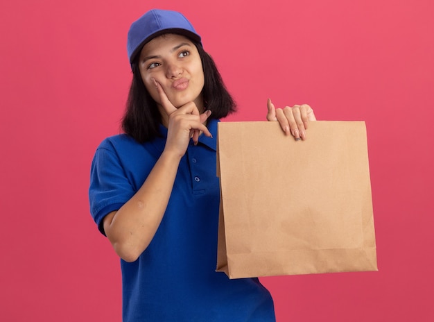 Jovem entregadora de uniforme azul e boné segurando um pacote de papel olhando para o lado perplexa em pé sobre a parede rosa