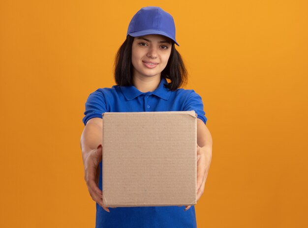 Jovem entregadora de uniforme azul e boné segurando uma caixa de papelão com um sorriso no rosto em pé sobre uma parede laranja