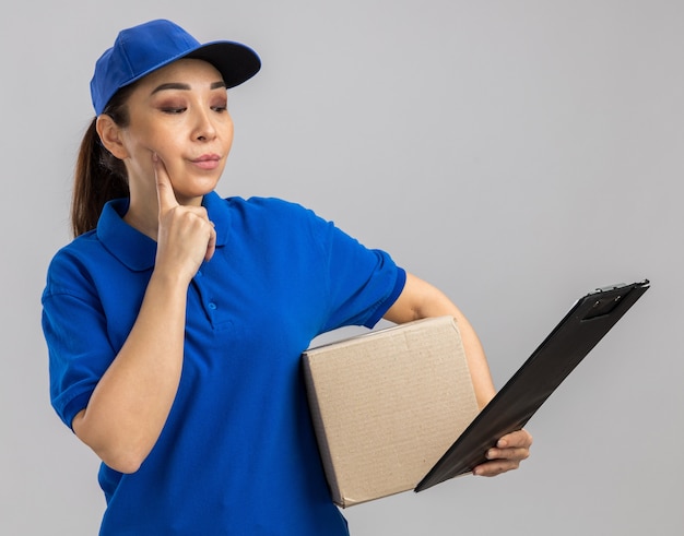 Jovem entregadora de uniforme azul e boné segurando uma caixa de papelão e uma prancheta olhando para ela com expressão pensativa e pensando em pé sobre uma parede branca