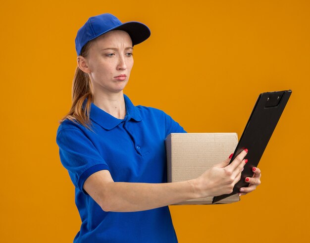 Jovem entregadora de uniforme azul e boné segurando uma caixa de papelão e uma prancheta olhando para ela com uma cara séria