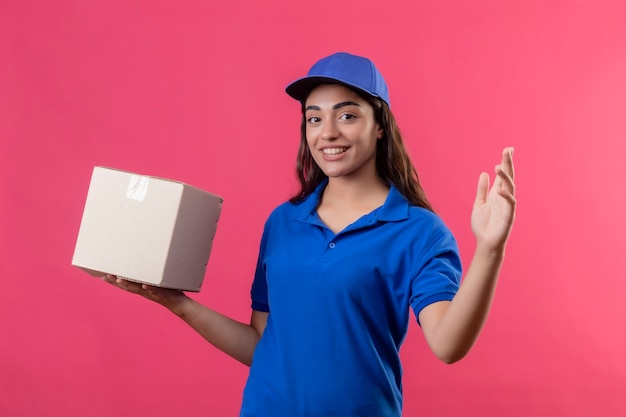 Jovem entregadora de uniforme azul e boné segurando uma caixa de papelão, levantando a mão, olhando para a câmera com um sorriso no rosto feliz e positivo em pé sobre um fundo rosa