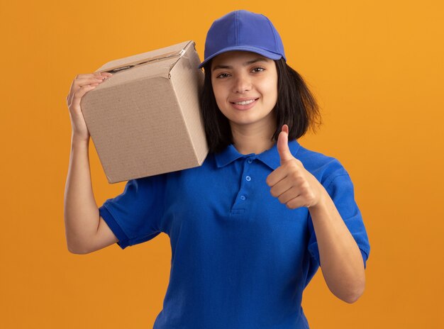 Jovem entregadora de uniforme azul e boné segurando uma caixa de papelão no ombro sorrindo e mostrando os polegares em pé sobre a parede laranja