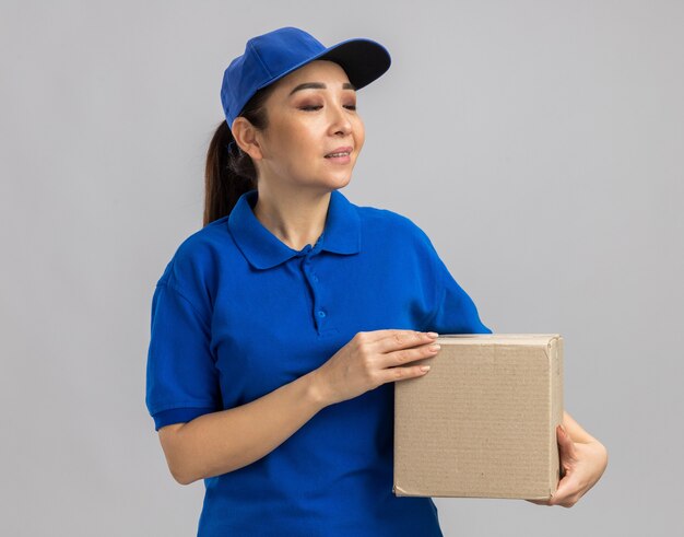 Jovem entregadora de uniforme azul e boné segurando uma caixa de papelão olhando para o lado com expressão confiante em pé sobre uma parede branca