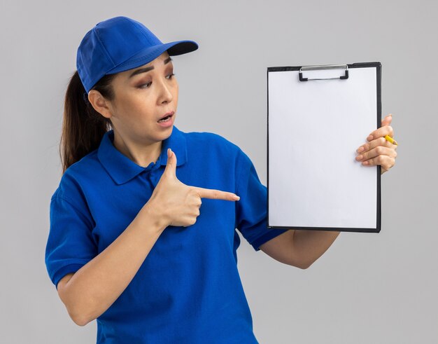 Jovem entregadora de uniforme azul e boné segurando uma prancheta com páginas em branco apontando com o dedo indicador para ela, surpresa em pé sobre uma parede branca