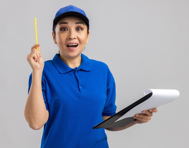 Jovem entregadora de uniforme azul e boné segurando uma prancheta e uma caneta feliz e surpresa