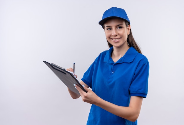 Jovem entregadora de uniforme azul e boné segurando uma prancheta e uma caneta olhando para o lado com um sorriso confiante no rosto