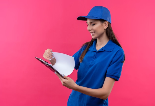 Foto grátis jovem entregadora de uniforme azul e boné segurando uma prancheta, olhando para páginas em branco com um sorriso no rosto