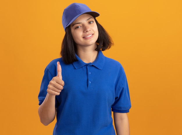 Jovem entregadora de uniforme azul e boné sorrindo amigavelmente mostrando os polegares em pé sobre uma parede laranja