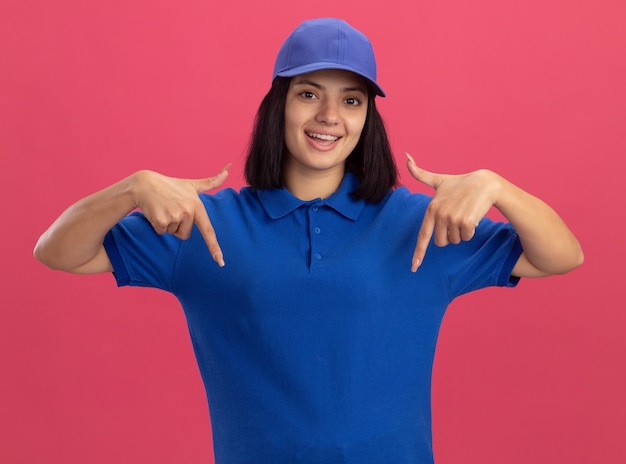 Jovem entregadora de uniforme azul e boné sorrindo, apontando com o dedo indicador para baixo, em pé sobre a parede rosa