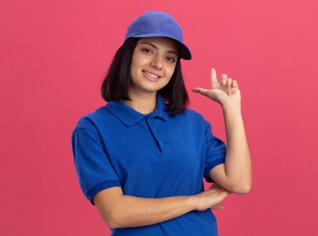 Jovem entregadora de uniforme azul e boné sorrindo, apontando com o indicador figner em pé sobre a parede rosa