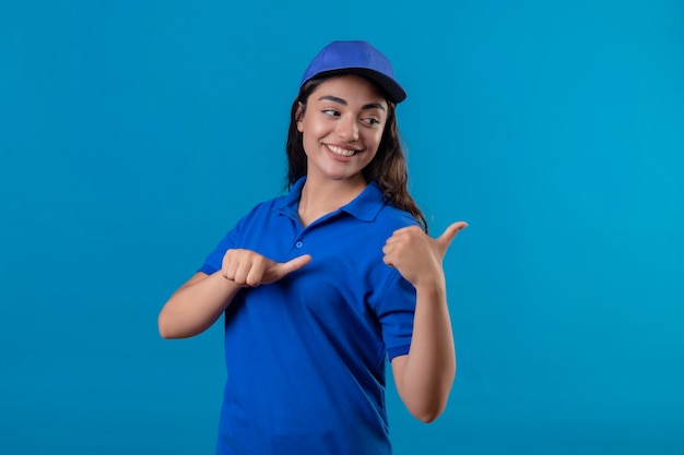 Foto grátis jovem entregadora de uniforme azul e boné sorrindo confiante apontando para o lado com os polegares em pé sobre um fundo azul