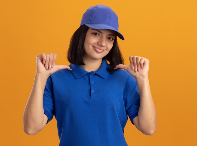 Jovem entregadora de uniforme azul e boné sorrindo confiante apontando para si mesma em pé sobre a parede laranja