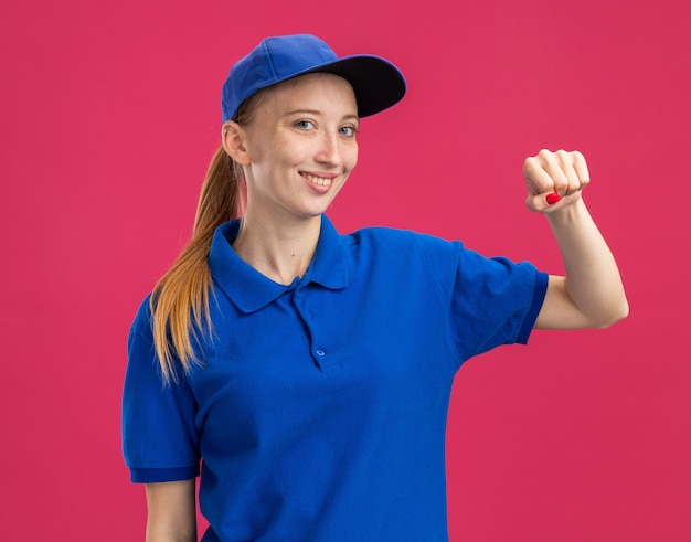 Jovem entregadora de uniforme azul e boné sorrindo, confiante, feliz e positiva, mostrando o punho em pé sobre a parede rosa