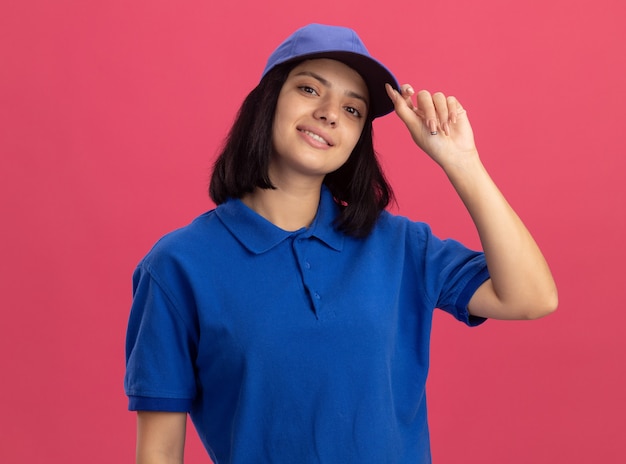 Jovem entregadora de uniforme azul e boné sorrindo confiante fixando o boné em pé sobre a parede rosa