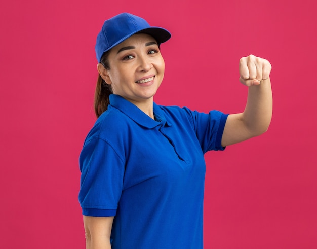 Foto grátis jovem entregadora de uniforme azul e boné sorrindo confiante levantando o punho