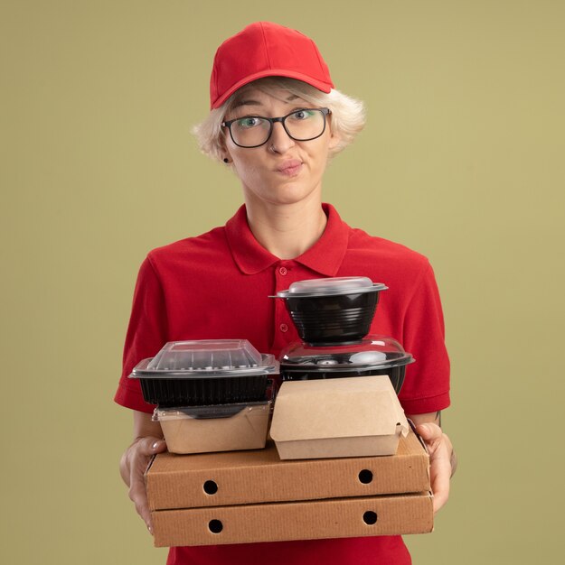 Jovem entregadora de uniforme vermelho e boné de óculos segurando caixas de pizza e pacotes de comida com expressão cética franzindo os lábios em pé sobre a parede verde