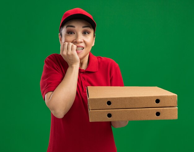 Jovem entregadora de uniforme vermelho e boné segurando caixas de pizza estressada e nervosa