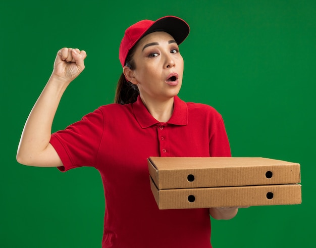 Jovem entregadora de uniforme vermelho e boné segurando caixas de pizza, levantando o punho e sendo surpreendida em pé sobre a parede verde