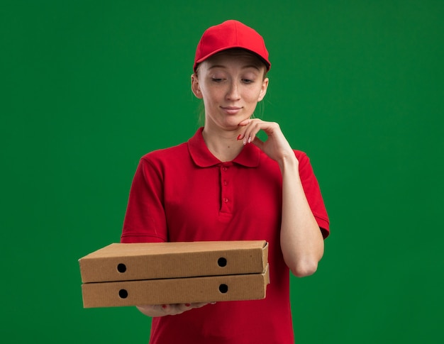 Jovem entregadora de uniforme vermelho e boné segurando caixas de pizza olhando para elas com a mão no queixo pensando em pé sobre a parede verde