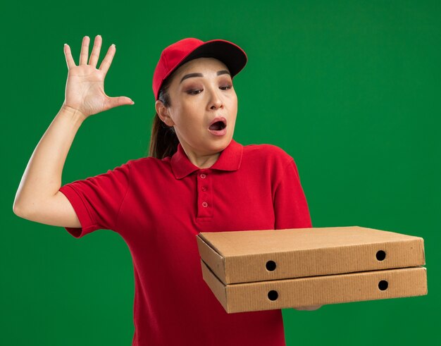 Jovem entregadora de uniforme vermelho e boné segurando caixas de pizza parecendo espantada e surpresa com o braço levantado em pé sobre a parede verde