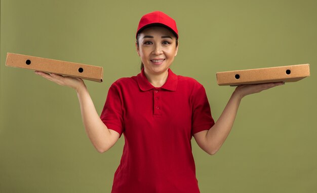 Jovem entregadora de uniforme vermelho e boné segurando caixas de pizza, sorrindo amigavelmente em pé sobre a parede verde