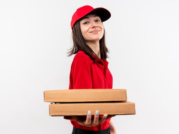 Jovem entregadora de uniforme vermelho e boné segurando caixas de pizza sorrindo com uma cara feliz em pé sobre uma parede branca Foto gratuita