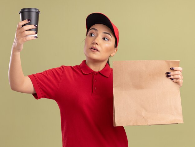 Jovem entregadora de uniforme vermelho e boné segurando um pacote de papel olhando para a xícara de café na outra mão com uma cara séria em pé sobre a parede verde