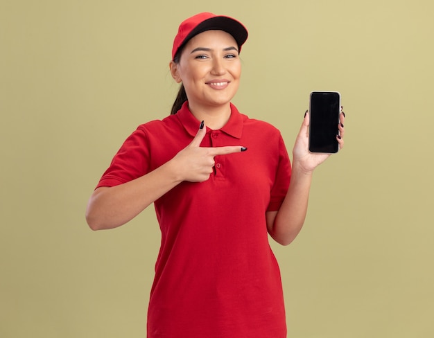 Jovem entregadora de uniforme vermelho e boné segurando um smartphone apontando com o dedo indicador para ele e sorrindo confiante em pé sobre a parede verde