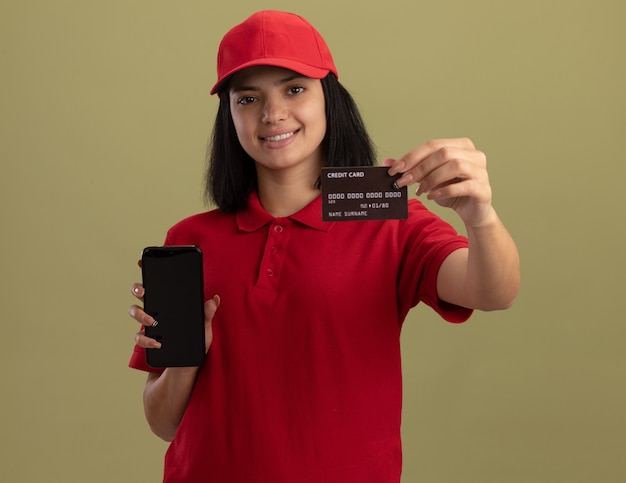 Jovem entregadora de uniforme vermelho e boné segurando um smartphone, mostrando o cartão de crédito sorrindo alegremente em pé sobre a parede de luz