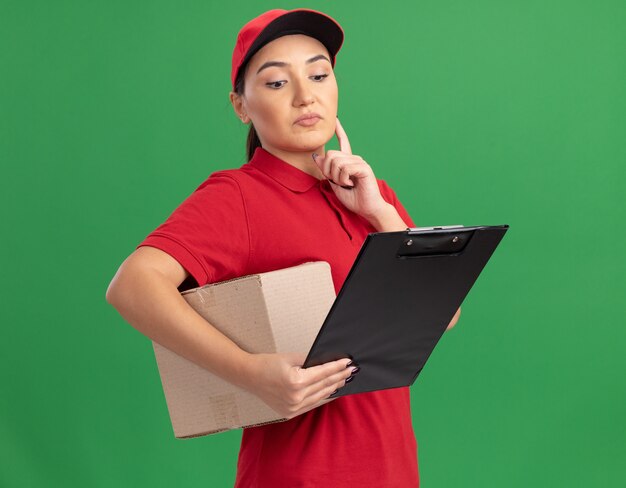 Jovem entregadora de uniforme vermelho e boné segurando uma caixa de papelão com uma prancheta olhando para ela com expressão pensativa em pé sobre a parede verde