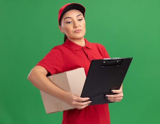 Jovem entregadora de uniforme vermelho e boné segurando uma caixa de papelão com uma prancheta olhando para ela com uma cara séria em pé sobre a parede verde