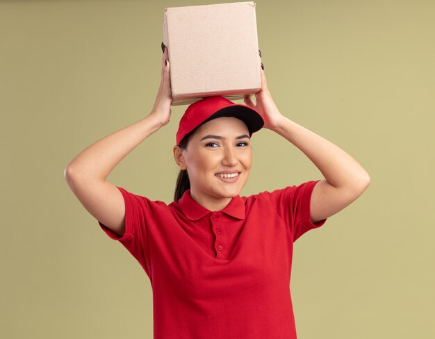 Jovem entregadora de uniforme vermelho e boné segurando uma caixa de papelão na cabeça, olhando para a frente, sorrindo alegremente em pé sobre a parede verde