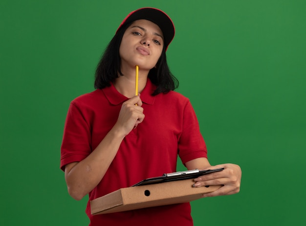 Jovem entregadora de uniforme vermelho e boné segurando uma caixa de pizza e uma prancheta com lápis perplexa em pé sobre a parede verde