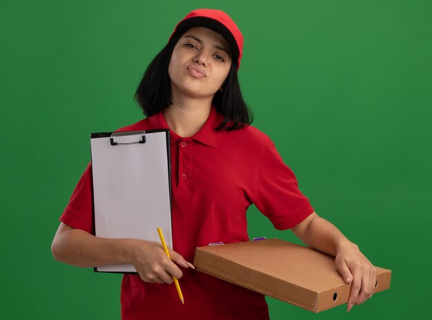 Jovem entregadora de uniforme vermelho e boné segurando uma caixa de pizza e uma prancheta com um lápis com expressão confiante em pé sobre a parede verde