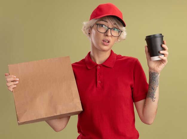 Jovem entregadora de uniforme vermelho e boné usando óculos, segurando um pacote de papel e uma xícara de café, confusa e muito ansiosa em pé sobre a parede verde