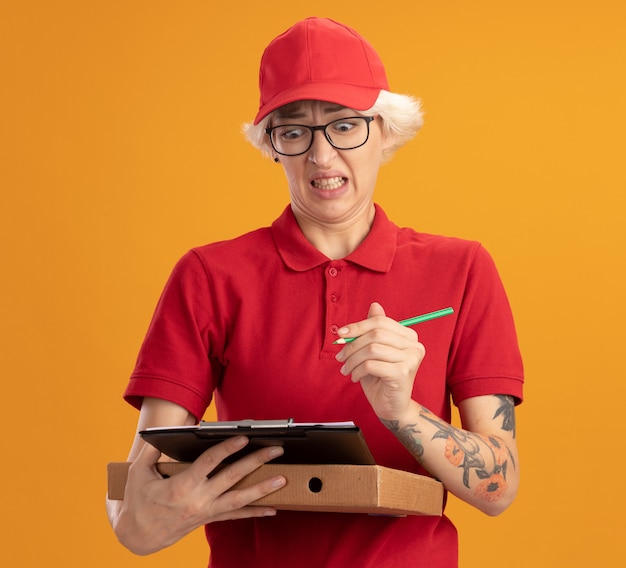 Jovem entregadora de uniforme vermelho e boné usando óculos, segurando uma caixa de pizza e uma prancheta com um lápis, parecendo confusa e desapontada em pé sobre uma parede laranja